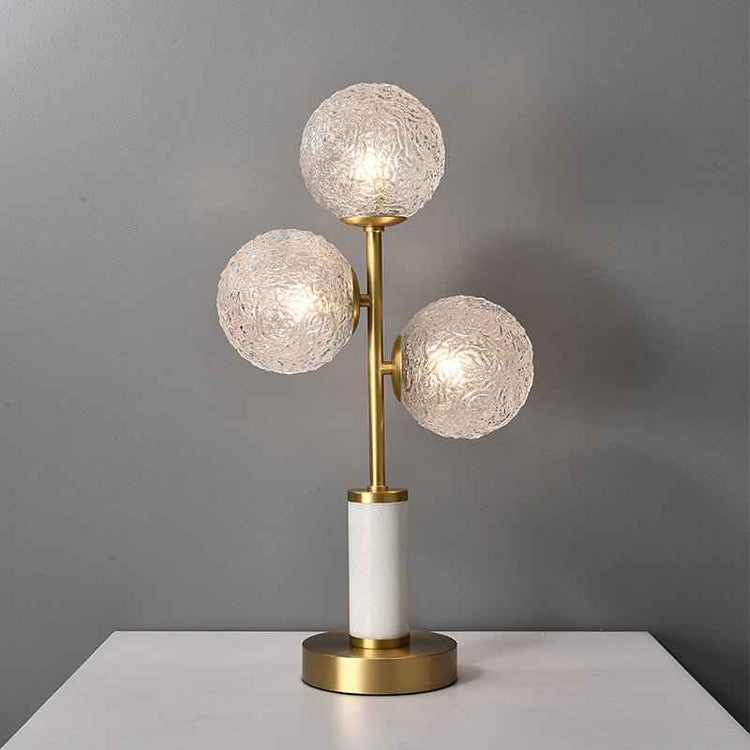 Lampe à poser 3 lumières : trois globes en verre texturé sur tige laiton, socle blanc et base dorée.