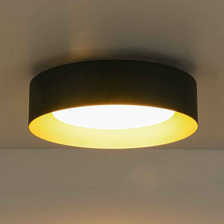 Plafonnier LED tambour noir, anneau doré et lumière chaude à travers verre laiteux.